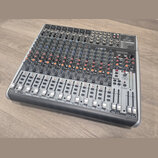 Stříbrný mixážní pult značky behringer s nápisem XENYX QX2222USB. Zařízení obsahuje numerousto potenciometrů v modré a oranžové barvě, posuvníky fade…