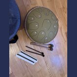Kruhlový steel tongue drum v olivové barvě s hnědým pleteným okrajem. Na povrchu jsou vyryté tóny s barevnými značkami a uprostřed je malý nápis "the…
