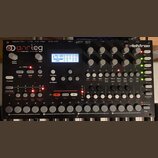 Černý analogový syntetizátor s bílými klávesami. Na horním panelu jsou nápisy analog a elektron, displej s modrým podsvícením a řada šedých otočných…
