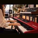 Červený digitální klavír s nápisem nord piano 5. Nástroj má bílé a černé klávesy a ovládací panel s několika knoflíky a displejem. V pozadí je umístě…