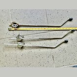 Tři kovové chromované ramena mikrofonních stativů s černými plastovými rukojtěmi. Vedle horního ramene je položen žlutý metr. Na kovových površích js…