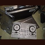 Bílý DJ kontrolér s nápisem TRAKTOR KONTROL S4. Vedle něj je položen hnědý karton s nápisem TRAKTOR KONTROL S4 a v pozadí černý transportní kufr s lo…
