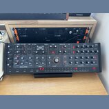 Černý analogový synthesizér s velkým množstvím ovládacích knoflíků a přepínačů. Na panelu jsou vidět nápisy OSCILLATORS, MIXER, FILTER a ENVELOPES. V…