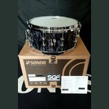 Tmavě modrý buben s chromovaným obručováním a ladicími šrouby. Nástroj stojí na hnědé papírové krabici s nápisy SONOR Made in Germany a SQ2 DRUM SYST…