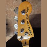 Fotografie zachycuje hlavu basové kytary z přírodního dřeva s čtyřmi chromovanými ladičkami. Na povrchu je černý nápis Squier BY FENDER a PRECISION B…