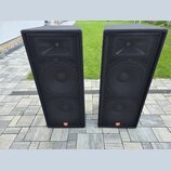 Dvě černé reproduktorové bedny s textilním potahem a kovovými mřížkami. Každý reproduktor má v dolní části oranžové logo JBL. Povrch textilu vykazuje…