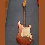 Elektrická kytara s růžovo-měděným lakem a bílým pickguardem. Na hlavě hředu je viditelné logo Fender. Lak na těle vykazuje známky opotřebení, zejmén…