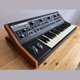 Klávesový syntetizátor s černým ovládacím panelem a dřevěným šasi. Na panelu je nápis „interstudio analog synthesizer“ a logo „moog“. Nástroj obsahuj…