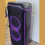 Černý party reproduktor s fialovým LED podsvícením. Na přední mřížce je umístěno zlaté logo JBL. Povrch je kombinací tkaniny a plastu s žebrovaným vz…