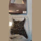 Sada předmontovaného pickguardu s mechanismy snímačů v průhledném plastovém obalu. Pickguard má černý mramorovaný vzor a tři černé snímače s bílým lo…