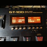 Černý efektový procesor s nápisem GT-100 COSM AMP EFFECTS PROCESSOR. Zařízení má dvě oranžové displeje s texty U01-4, Solo a dalšími zkratkami efektů…
