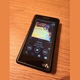 Černý digitální přehrávač značky SONY s displejem. Na obrazovce je zobrazen album cover Norah Jones, název skladby Sunrise a album Feels Like Home. V…