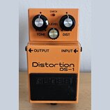 Oranžový kytarový efekt s nápisy Distortion a DS-1. Na horní části jsou tři ovládací knoflíky s popisky TONE, LEVEL a DIST a kontrolka CHECK. Vprostř…