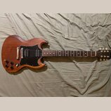 Elektrická kytara s hnědým dřevěným povrchem a černým pickguardem. Na hlavě je viditelné logo Gibson. Kytara má dva humbuckery, čtyři ovládací knoflí…