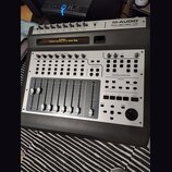 Stříbrno-černý kontrolní povrch M-AUDIO PROJECTMIX I/O. Na displeji je nápis „M-Audio Control Surface is in Sonar Mode“. Zařízení obsahuje faderové p…