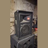 Černý reproduktor v bedně potažené tmavým kobercem s kovovými hranami. V horní části je rohové hrdlo s oranžovým logem JBL, ve spodní části je kruhov…