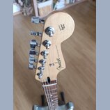Záběr na hlavu elektrické kytary z přírodního dřeva s chromovanými ladičkami. Na zadní straně jsou nápisy Fender, STRATOCASTER® a ORIGINAL Contour Bo…