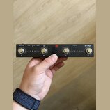 Černý obdélníkový přístroj s kovovým povrchem a čtyřmi konektory. Na horní straně jsou nápisy (U O H), USB, HOST a PEDAL, v pravém rohu je logo M-VAV…