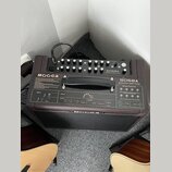 Hnědý akustický zesilovač s nápisem MOOGER a označením modelu SDSØA ACOUSTIC AMPLIFIER. Horní panel obsahuje ovládací knoflíky, přepínače a vstupy, z…