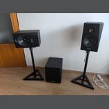 Sada černých reproduktorů tvořená dvěma regálovými boxy na stojanách a jedním subwooferem. Na přední straně všech tří komponentů je viditelné logo AR…