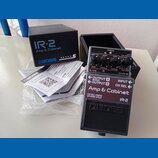 Na fotografii je kytarový pedál Boss IR-2 Amp & Cabinet v modré barvě s černým spodním panelem. Vedle pedálu je umístěna originální černá krabice s n…