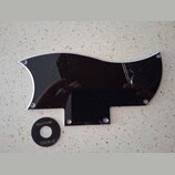 Černý plastový plech ochranný (pickguard) s vyleštěným stříbrným okrajem a otvory pro šrouby. Vedle něj je umístěn malý černý kruhový přepínač s bílý…