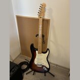 Elektrická kytara s tłem v barvě sunburst a bílým pickguardem. Na hlavě hředu je viditelné logo Fender. Kytara stojí na černém stojanu před korkovou…