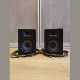 Dva černé studiové reproduktory umístěné vedle sebe na dřevěné podlaze. Každý reproduktor má v horní části výškoveý reproduktor a ve spodní části bas…