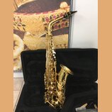 Zlatý saxofon umístěný v otevřeném černém kufru. Na zvonu nástroje je viditelné logo a nápis "YAMAHA". Povrch instrumentu je lesklý, s bílými odpoutá…