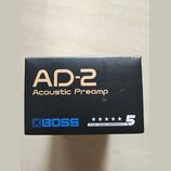 Černá krabice s produktem Boss AD-2. Na přední straně je zlatým písmem nápis AD-2 Acoustic Preamp a modré logo BOSS. V pravém dolním rohu je bílý náp…