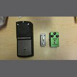 Na světlém dřevěném povrchu leží tři kytarové efekty. Zleva je zelený pedál s nápisy MXR a GT-100, vybavený dvěma otočnými knoflíky označenými jako G…