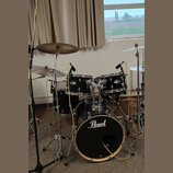 Černá bicí souprava značky Pearl s kovovým hardwarem. Sestává z basového bubnu, dvou tomů a jednoho floortomu, doplněných o tři činely. Na basovém bu…