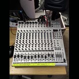 Stříbrný mixážní pult s více kanály, vybavený řadou potenciometrů a posuvníků. V horní části je nápis BEHRINGER UX2044UP-USB. Na spodní hraně je nale…