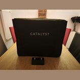 Černý textilní kryt na hudební zesilovač s bílým nápisem CATALYST uprostřed. Před krytem leží černý stupňovací spínací pedál s nápisy CATALYST, CHANN…
