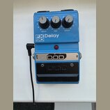 Modrý kytarový efekt s třemi ovládacími knoflíky popsanými jako DELAY, MIX a REPEAT. Na přední straně je nápis FX 90 Delay a červená kontrolka. Spodn…