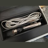 V černé přepravce s polstrováním se nachází mikrofon a bílý kabel. Mikrofon má stříbrný kovový trup, černou horní část s mřížkou a v dolní části je v…