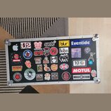 Černý transportní kufr s hliníkovými hranami a kovovými rohy, který je polepen mnoha nálepkami. Mezi viditelnými nápisy a logy jsou: Apple, U2, RADIO…