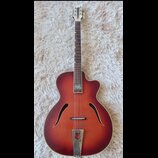 Kytara s polodubnovým tělem v červenohnědém sunburst provedení. Na hmatníku je viditelný světlý nápis, který není čitelně rozlučitelný. Nástroj vykaz…