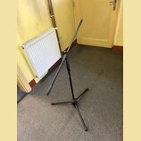 Černý kovový stativ s rozkládacím trojnohým základem a teleskopickým ramenem. Na jedné z nohou je viditelný svislý nápis "TAMA". Povrch stativu je ma…