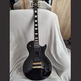 Černá elektrická kytara s tvarem singlecut a bílým lemováním hran. Na hlavě je nápis Epiphone, hриф má vložky z perlece a kytara je vybavena dvěma če…