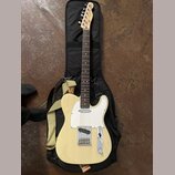 Krémová elektrická kytara typu Telecaster s bílým pickguardem, položená na černém softcasu. Na hlavě hředu je viditelné logo Fender. Kytara má žlutý…