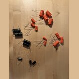 Na dřevěném povrchu je rozprostřeno několik elektronických součástek, konkrétně kondenzátorů. Většina z nich má oranžový povrch a kovové vodiče, osta…