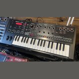 Černý syntezátor s klaviatura značky Roland, model JD-Xi. Horní panel obsahuje červený displej, početné ovládací knofíky a tlačítka rozdělená do sekc…
