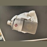 Stříbrný kondenzátorový mikrofon s kovovou síťkovou hlavou, položený na bílé látce. Na těle je vidět logo v červeném kosočtverci a nápis NEUMANN MICR…