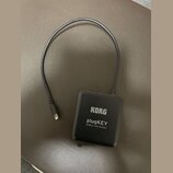 Černý obdélníkový adaptér s připojeným kabelem a konektorem. Na povrchu je bílý nápis KORG, plugKEY a MOBILE MIDI/AUDIO. Povrch zařízení je matný a v…