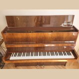 Hnědé dřevěné piano s lakovaným povrchem. Na čelní straně nad klávesami je zlatý nápis PETROF. V otevřeném horním víku jsou vidět struny a v pravé čá…