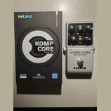 Na fotografii je stříbrný efektový pedál s černými ovládacími knoflíky a přepínačem. Na pedálu jsou nápisy KOMP CORE DELUXE MKII a logo NUX. Vedle pe…