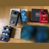 Fotografie zobrazuje čtyři kytarové efekty s jejich krabicemi na dřevěné podlaze. Jsou zde pedály: černý tc electronic DITTO STEREO LOOPER, modrý ANG…