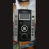 Stříbrný perkusní modul značky Roland s černým předním panelem. Na horní části je nápis PERCUSSION SOUND MODULE TD-9 a v dolní části logo V-Drums. Mo…