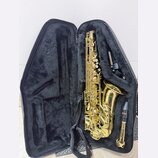 Zlatý saxofon v černém polstrovaném kufru. Na zvonu nástroje je nápis "YAMAHA". V kufru je vedle saxofonu umístěn munčník s nápisem "YAMAHA" a černý…